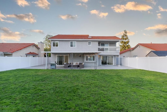 back house at dusk with a patio area, a balcony, and a lawn