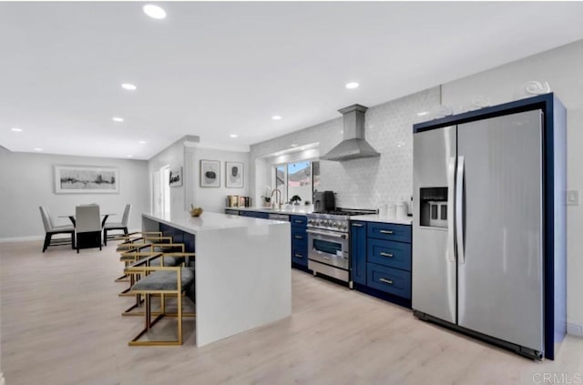 kitchen featuring blue cabinets, a breakfast bar, a center island, stainless steel appliances, and wall chimney range hood