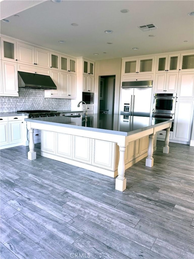 kitchen with a kitchen island with sink, a kitchen breakfast bar, built in appliances, decorative backsplash, and exhaust hood