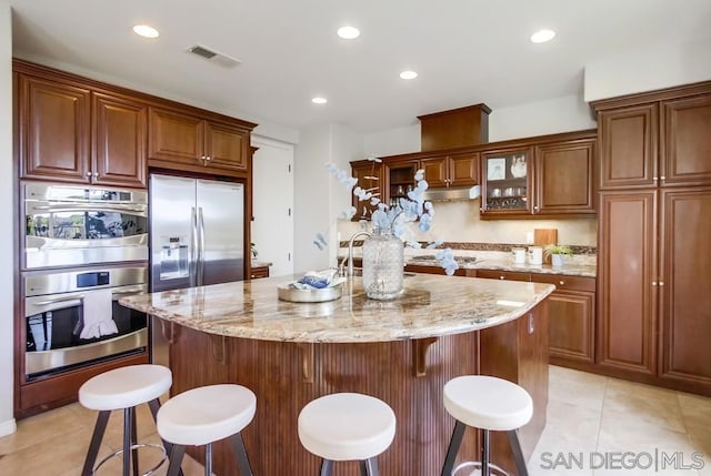 kitchen with light tile patterned floors, appliances with stainless steel finishes, a center island with sink, light stone countertops, and a kitchen bar