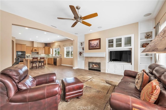 living room with built in shelves, a tile fireplace, light hardwood / wood-style floors, and ceiling fan