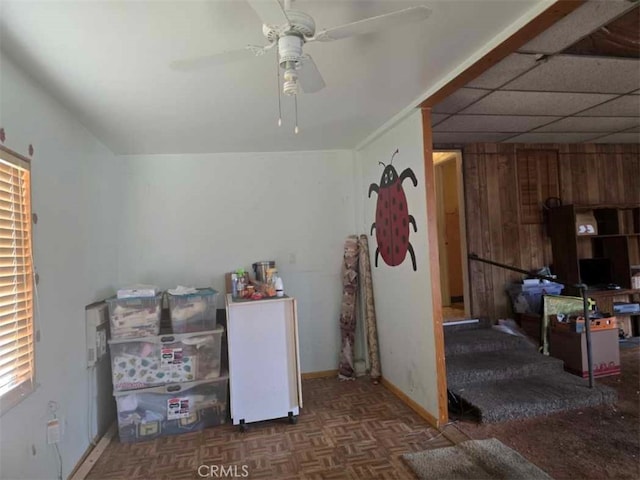 interior space featuring a drop ceiling, a ceiling fan, baseboards, and wood walls