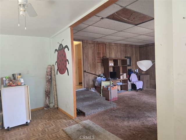 below grade area with a paneled ceiling, baseboards, wood walls, and ceiling fan