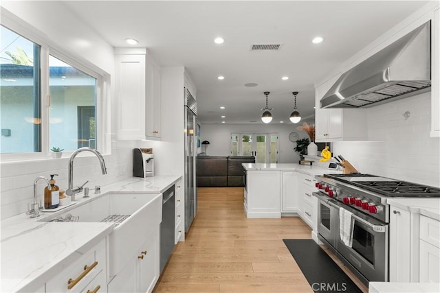 kitchen with hanging light fixtures, high quality appliances, light stone counters, white cabinets, and wall chimney exhaust hood