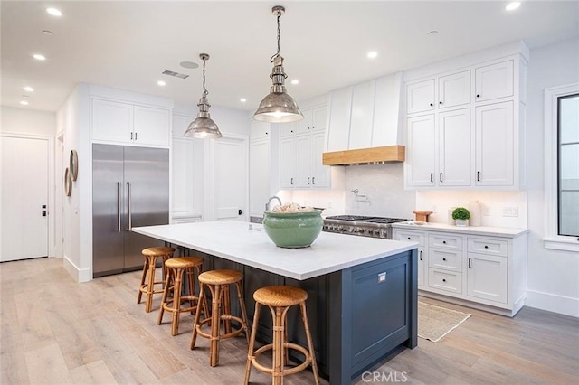 kitchen with premium range hood, hanging light fixtures, an island with sink, built in fridge, and white cabinets