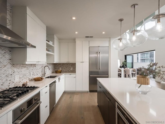 kitchen featuring appliances with stainless steel finishes, tasteful backsplash, white cabinets, hanging light fixtures, and wall chimney exhaust hood