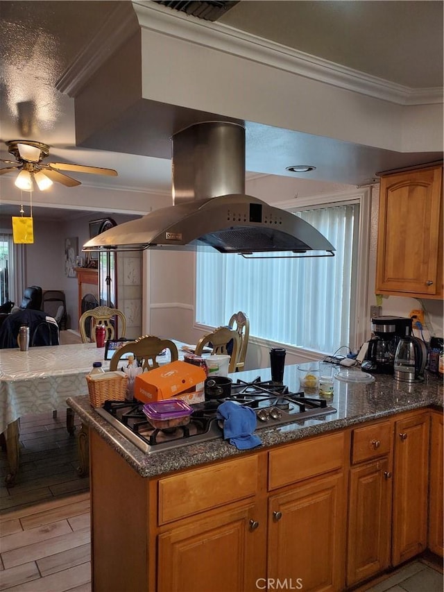 kitchen with island exhaust hood, ornamental molding, ceiling fan, dark stone counters, and light hardwood / wood-style floors