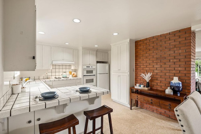 kitchen with white cabinetry, tile counters, a kitchen breakfast bar, white appliances, and brick wall