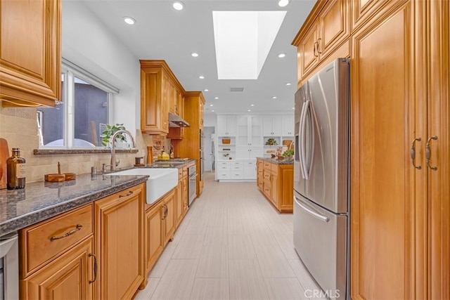 kitchen with sink, a skylight, dark stone countertops, stainless steel fridge, and backsplash