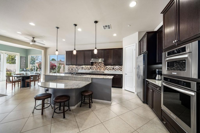 kitchen with pendant lighting, a breakfast bar, appliances with stainless steel finishes, light stone counters, and a center island with sink