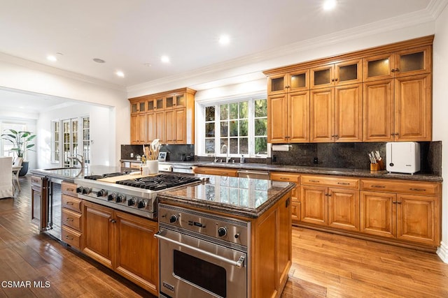 kitchen with appliances with stainless steel finishes, dark stone counters, crown molding, a center island with sink, and light wood-type flooring