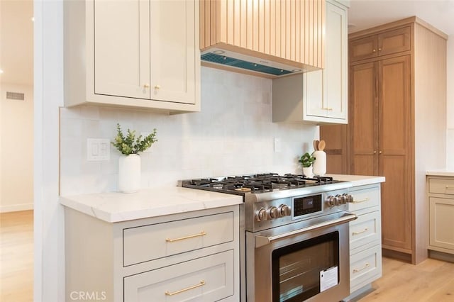 kitchen with backsplash, high end range, light stone counters, wall chimney range hood, and light hardwood / wood-style flooring