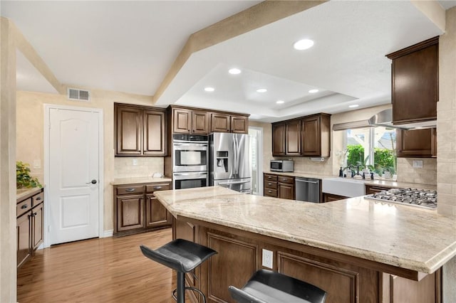 kitchen with a breakfast bar, appliances with stainless steel finishes, light stone counters, a raised ceiling, and light wood-type flooring