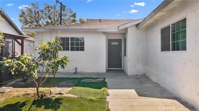 property entrance featuring stucco siding, a shingled roof, and a patio