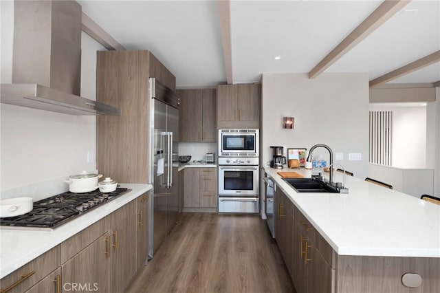 kitchen with beam ceiling, a sink, wall chimney range hood, built in appliances, and a peninsula