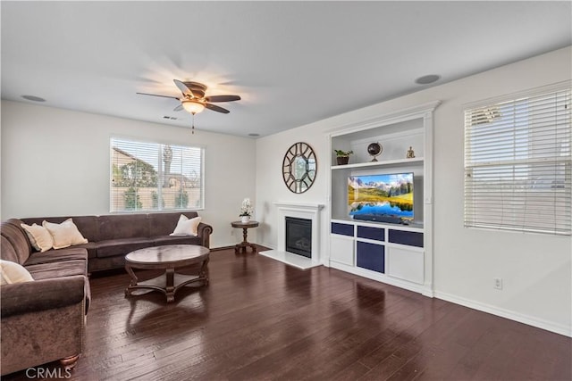 living room with built in shelves, a glass covered fireplace, wood finished floors, and a ceiling fan