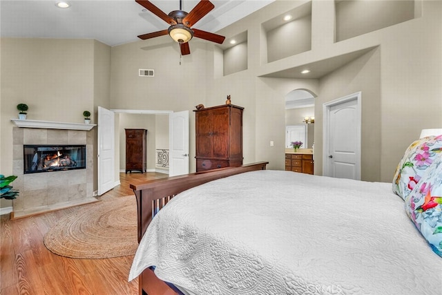 bedroom featuring hardwood / wood-style flooring, a tile fireplace, ceiling fan, a towering ceiling, and connected bathroom