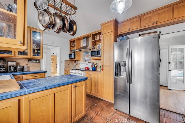 kitchen featuring tile countertops, appliances with stainless steel finishes, glass insert cabinets, tile patterned flooring, and wall chimney range hood