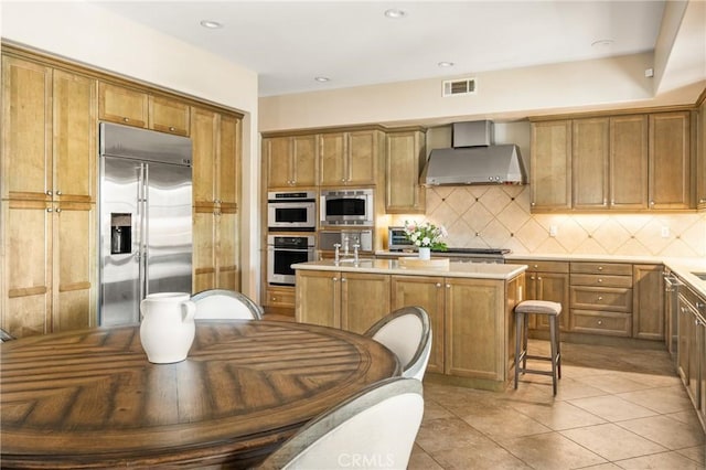 kitchen featuring light tile patterned flooring, built in appliances, a center island with sink, decorative backsplash, and wall chimney range hood