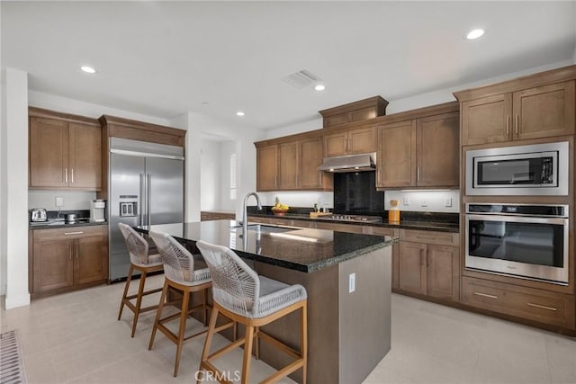 kitchen with an island with sink, sink, dark stone countertops, a kitchen bar, and built in appliances