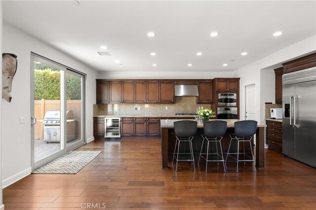 kitchen with a breakfast bar, a center island, ventilation hood, appliances with stainless steel finishes, and beverage cooler