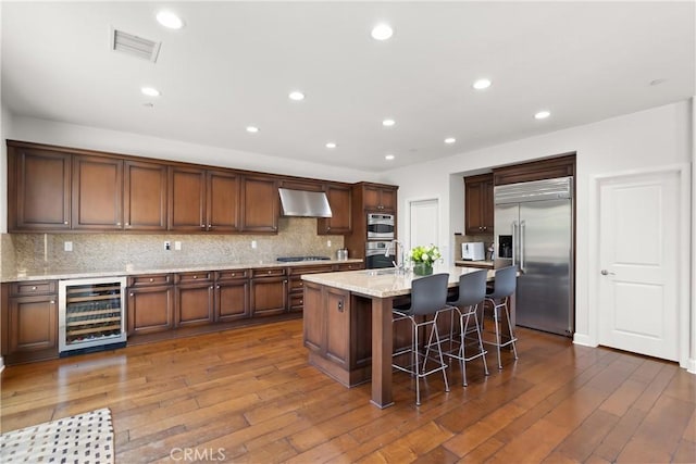 kitchen featuring stainless steel appliances, a kitchen breakfast bar, range hood, wine cooler, and a center island with sink