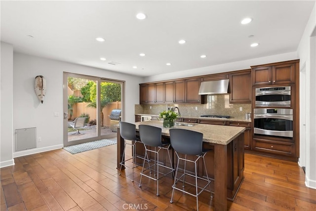 kitchen featuring stainless steel gas stovetop, backsplash, a kitchen breakfast bar, a kitchen island with sink, and wall chimney exhaust hood