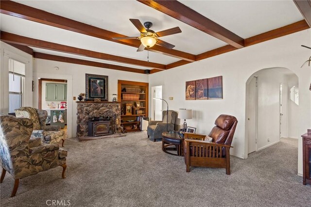 carpeted living room featuring arched walkways, a ceiling fan, beamed ceiling, a wood stove, and built in shelves