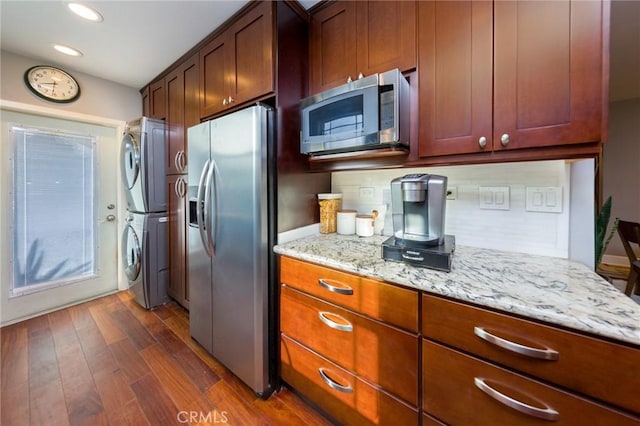 kitchen with light stone counters, appliances with stainless steel finishes, dark hardwood / wood-style flooring, stacked washing maching and dryer, and backsplash