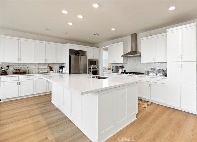 kitchen featuring wall chimney range hood, sink, a kitchen island with sink, stainless steel appliances, and white cabinets