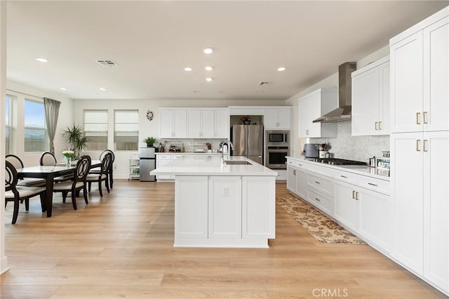 kitchen with appliances with stainless steel finishes, white cabinets, a kitchen island with sink, wall chimney range hood, and light wood-type flooring