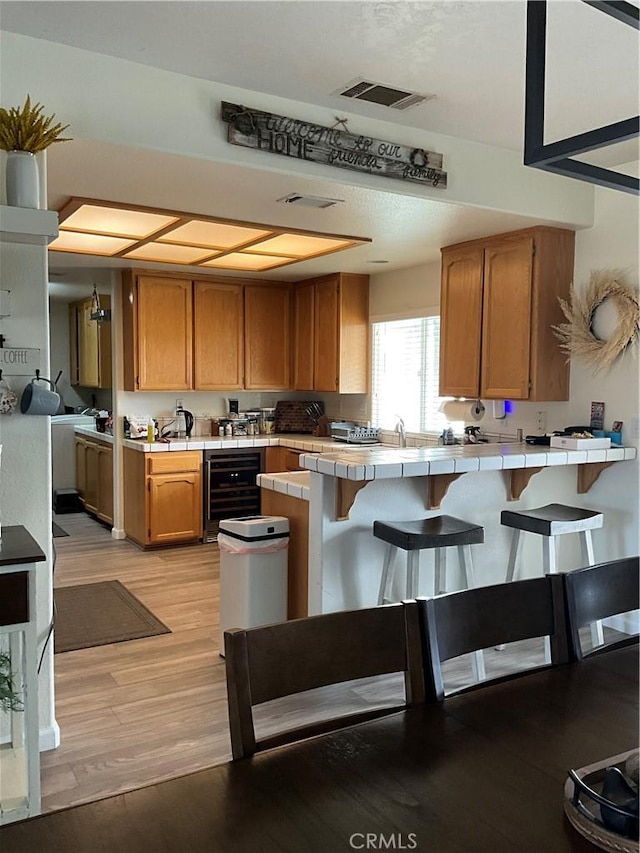 kitchen featuring a kitchen breakfast bar, tile counters, kitchen peninsula, beverage cooler, and light wood-type flooring