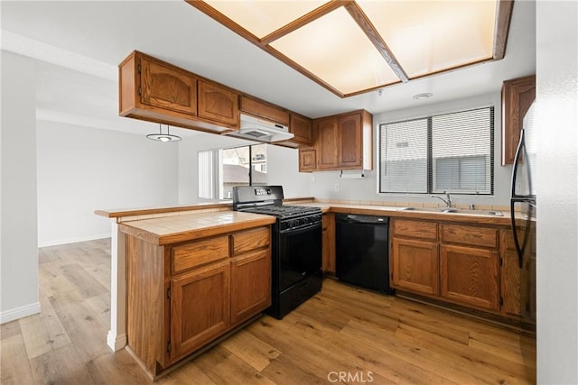 kitchen featuring brown cabinetry, a sink, a peninsula, under cabinet range hood, and black appliances