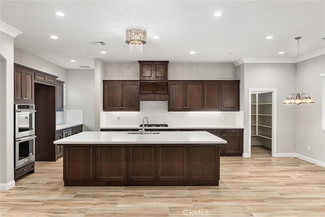 kitchen with dark brown cabinetry, hanging light fixtures, a kitchen island with sink, stainless steel double oven, and light hardwood / wood-style floors