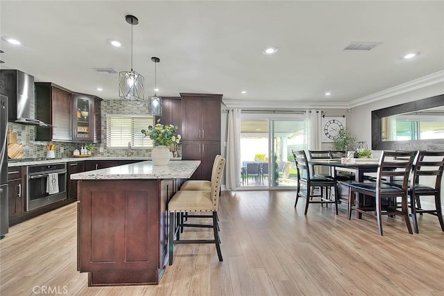 kitchen featuring wall chimney exhaust hood, crown molding, stainless steel oven, a kitchen island, and pendant lighting