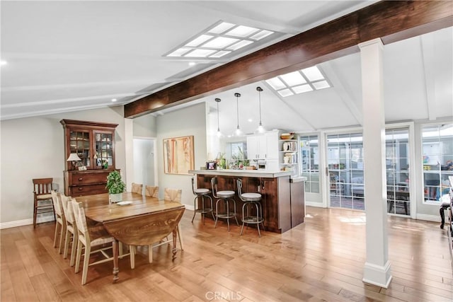 dining space with ornate columns, vaulted ceiling with skylight, bar area, and light wood-type flooring