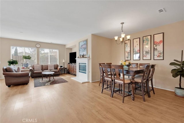 dining room featuring baseboards, visible vents, a glass covered fireplace, light wood-type flooring, and a notable chandelier