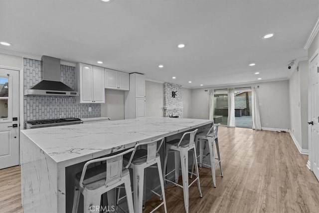 kitchen featuring white cabinetry, crown molding, wall chimney range hood, and light stone counters