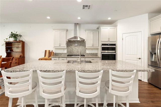 kitchen with white cabinetry, stainless steel appliances, a kitchen island with sink, decorative backsplash, and wall chimney range hood