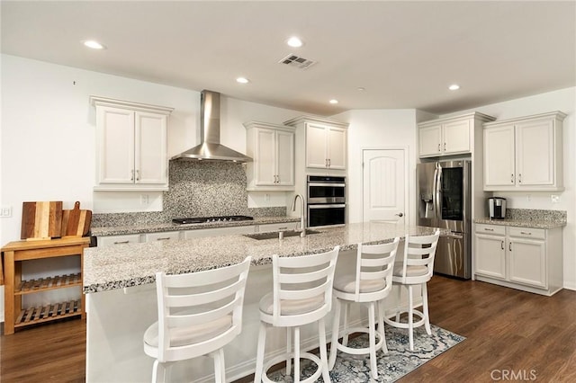 kitchen with sink, appliances with stainless steel finishes, a kitchen island with sink, light stone counters, and wall chimney exhaust hood