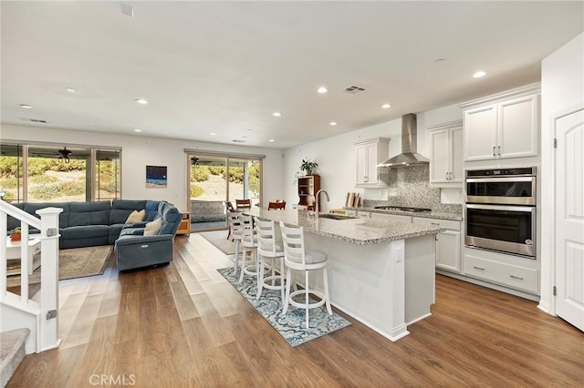 kitchen featuring a breakfast bar, a kitchen island with sink, stainless steel appliances, white cabinets, and wall chimney exhaust hood