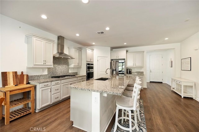 kitchen featuring sink, appliances with stainless steel finishes, an island with sink, light stone countertops, and wall chimney range hood