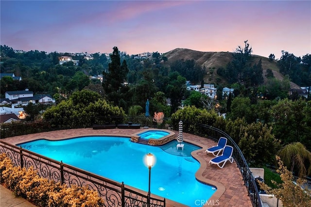 pool at dusk featuring fence, a mountain view, a pool with connected hot tub, and a patio