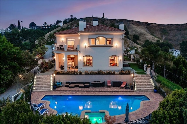 back of house at dusk with stairs, a tile roof, stucco siding, a chimney, and a balcony