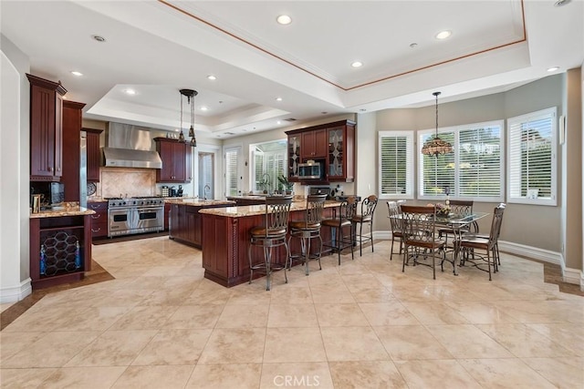 kitchen with a peninsula, baseboards, wall chimney exhaust hood, a tray ceiling, and stainless steel appliances