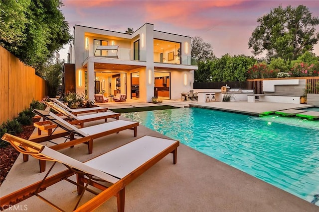 back house at dusk with a fenced in pool, exterior kitchen, a balcony, and a patio area