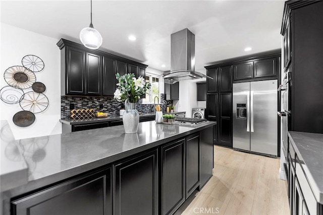 kitchen featuring appliances with stainless steel finishes, hanging light fixtures, tasteful backsplash, island exhaust hood, and light wood-type flooring