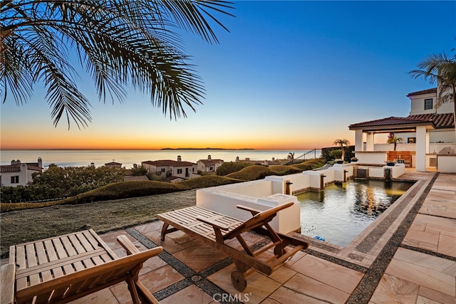 pool at dusk featuring a water view, a patio, and area for grilling