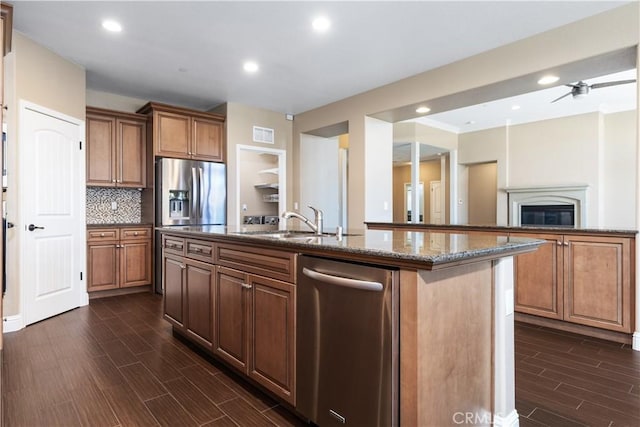 kitchen with sink, appliances with stainless steel finishes, dark stone countertops, backsplash, and a center island with sink