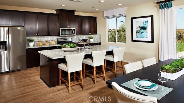 kitchen with appliances with stainless steel finishes, an island with sink, a breakfast bar area, dark brown cabinetry, and light stone countertops